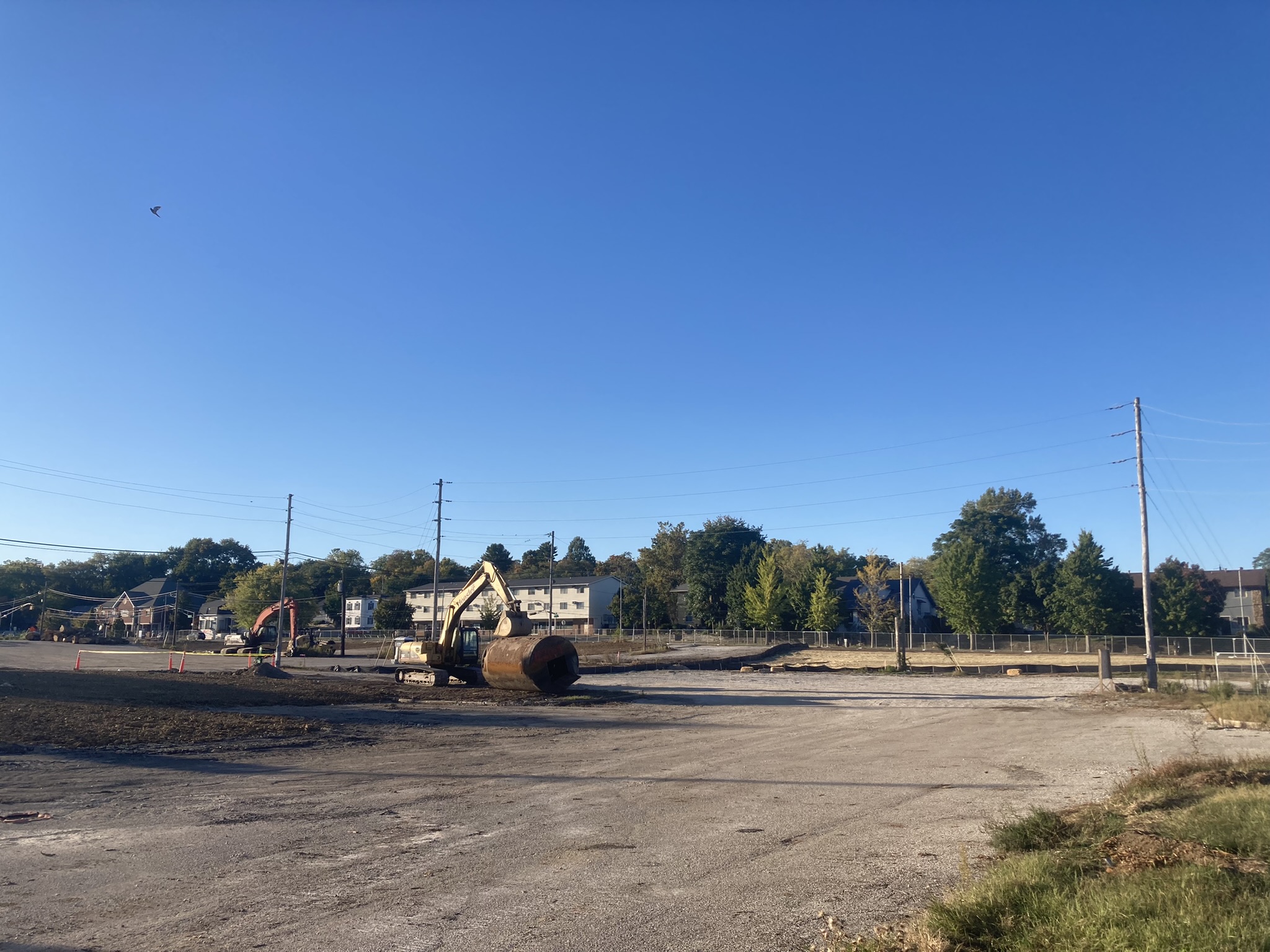 An excavator sits in a cleared, fenced, construction area; its bucket rests on a cylindrical tank. In the background are telephone poles and wires, trees, and residential (or small office) buildings. 