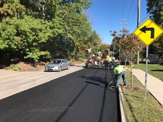 A City worker near a left turn road sign smoothes newly laid pavement. A silver sedan and City equipment are in the background. 