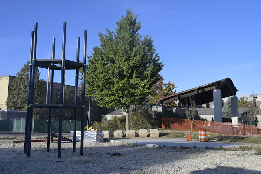 In the foreground is a newly installed two-level six-sided climber. In the background are a tree and the outdoor park stage, protected by construction net fencing. 