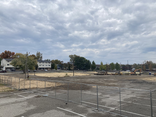 Image shows an excavator sits in a cleared, fenced, construction area. In the background are telephone poles and wires, trees, and residential (or small office) buildings. 