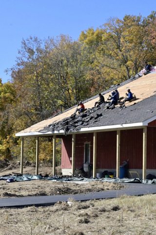 Photo shows a red barn at Goat Farm Park, with six people sitting on the roof and scraping off black shingles to reveal light colored decking beneath.