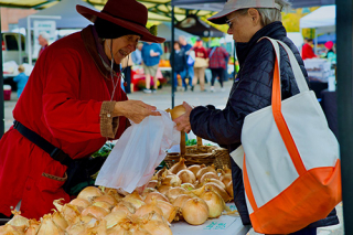 A woman farm vendor places a whole yellow onion into a plastic shopping bag for a Market customer. Photo by Merrill Hatlen.